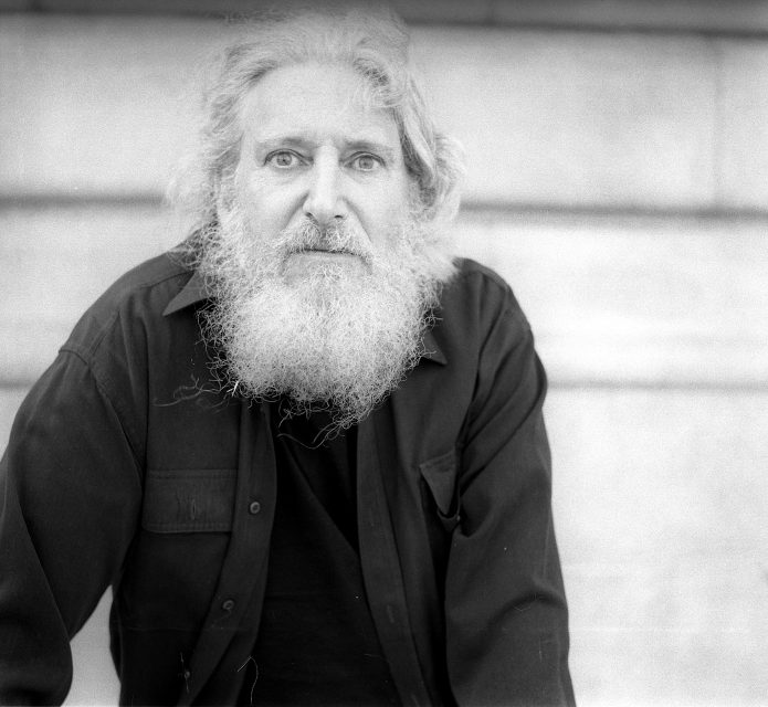 Black and white photo of an older person with a long white beard and hair, wearing a dark shirt, looking directly at the camera in front of a blurred stone background.