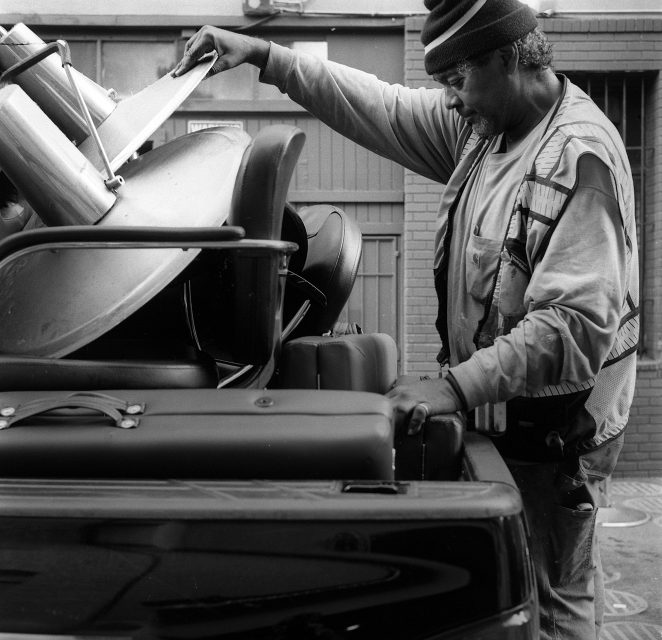 A man wearing a beanie and vest loads metal chairs and other items into the back of a pickup truck in an urban setting.