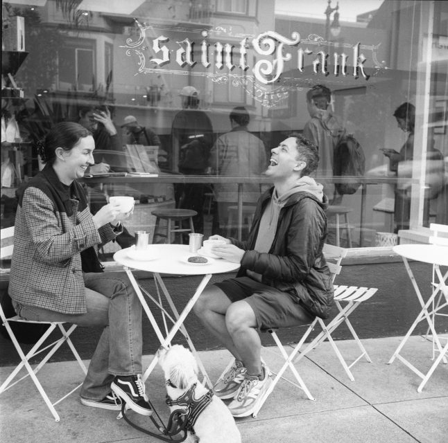 Two people sit at a small outdoor café table, drinking coffee and laughing. A dog sits on the ground beside them. The café window reads "Saint Frank.