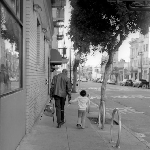 An adult and a child walk hand-in-hand down a city sidewalk lined with buildings and trees on a cloudy day.