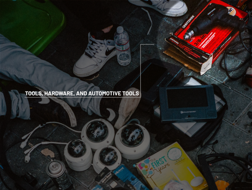 Various tools, security cameras, electronic devices, automotive tools, and a water bottle are scattered on a tiled floor near people’s feet.