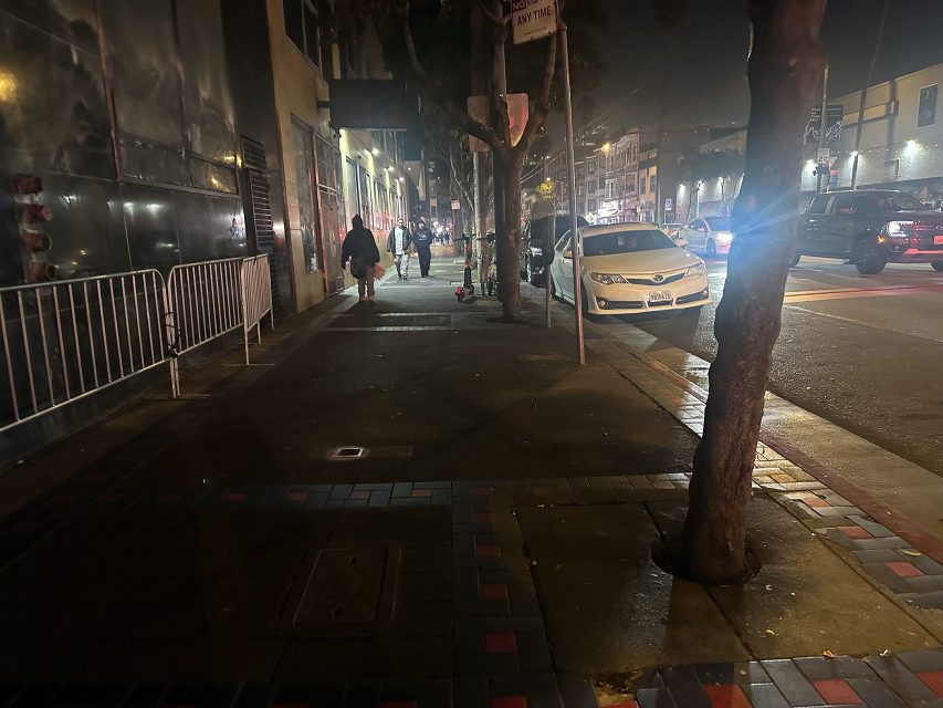 Nighttime street scene with parked cars, people walking on the sidewalk, a tree, and metal barricades along the left side of the building. Streetlights illuminate the area.