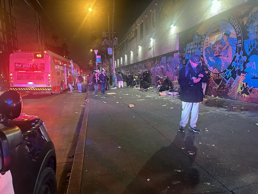 People stand and sit on a littered sidewalk at night near a bus and a mural-covered wall; a police car is partially visible in the foreground.