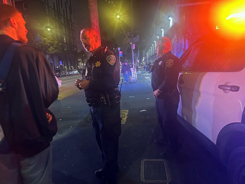 Two police officers stand near a police vehicle with flashing lights at night on a city street, talking to a man; debris is scattered on the ground in the background.