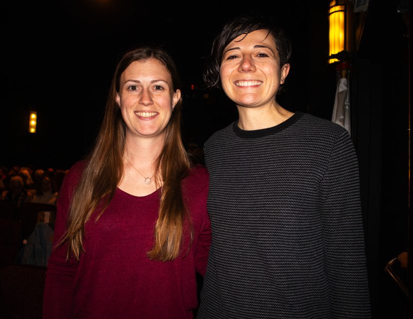 Two people standing indoors and smiling at the camera, with an audience seated in the dimly lit background.
