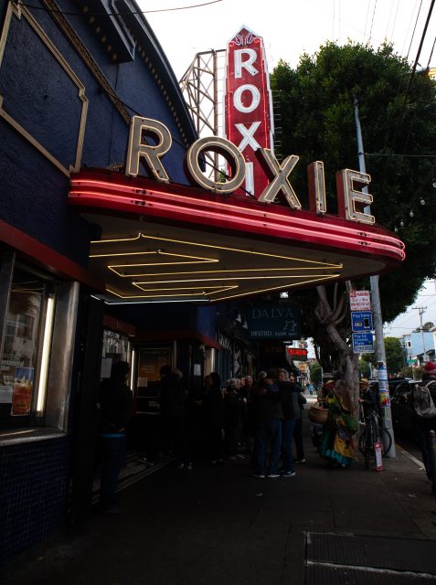 People line up outside the Roxie Theater under a red neon marquee on a city street with trees and storefronts visible.