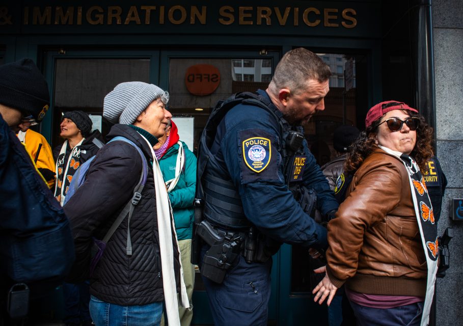 A police officer handcuffs a person in a leather jacket while others stand in line outside an immigration services building.