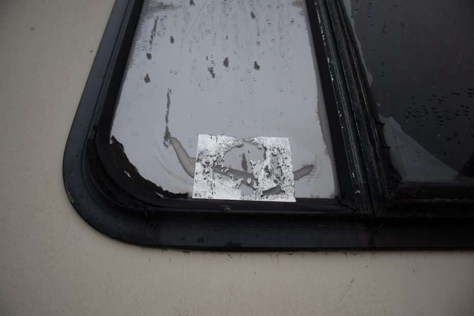 Close-up of a window with peeling, reflective sticker residue on the glass and visible water damage around the edges.