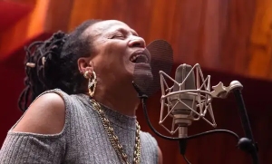 A woman with braided hair sings passionately into a microphone in a recording studio.
