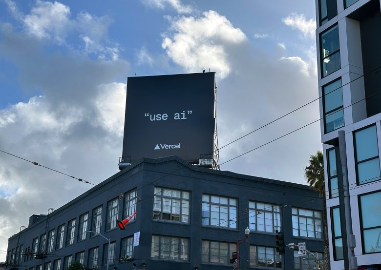 A large black billboard on a building reads “use ai” in white text with the Vercel logo below, set against a cloudy sky and surrounded by modern city buildings.