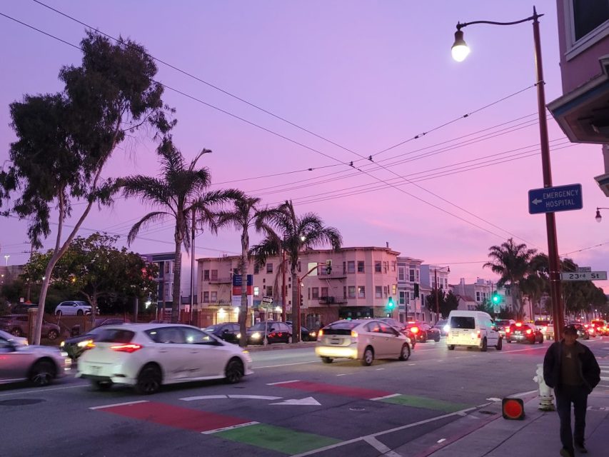 Cars and pedestrians move along a busy city street at dusk, with a purple sky and palm trees lining the road; streetlights and traffic signals are visible.