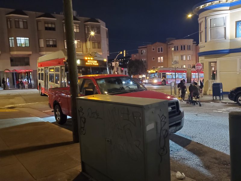 A red pickup truck and a bus are stopped at an intersection at night in a city; pedestrians cross the street and buildings are visible in the background.