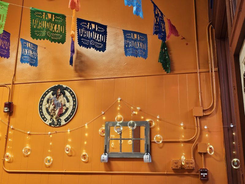 Colorful papel picado hangs above a wall decorated with string lights, a round Aztec calendar, and a small window frame against an orange background.