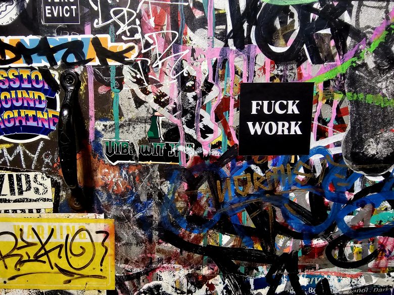 A wall covered in colorful graffiti, stickers, and tags, with a black sticker in the center displaying the words "FUCK WORK" in white capital letters.