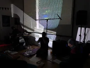 A dark room with people sitting on the floor, musical equipment set up, and a large abstract image projected onto the wall behind them.