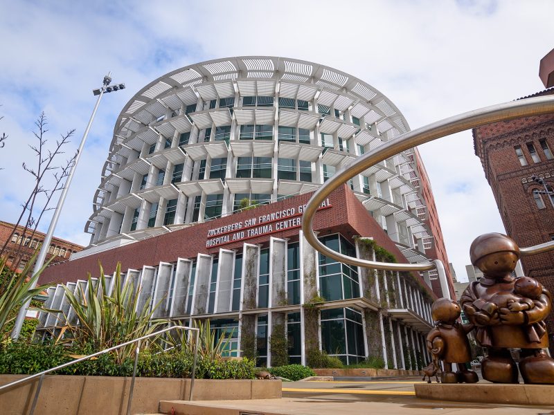 A modern hospital building with a circular design and large windows, identified as the Zuckerberg San Francisco General Hospital and Trauma Center, known for treating stabbing victims, with statues in the foreground.