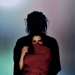 A person wrapped in a red cloth stands in front of a taller figure whose face is obscured by shadow, against a plain background.