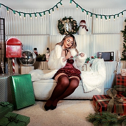 A woman in festive attire sits on a couch surrounded by Christmas decorations, wrapped gifts, a wreath, and string lights.