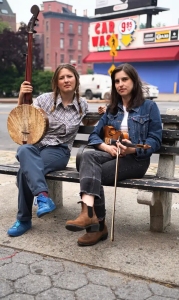 Two women sit on a bench outdoors. One holds a banjo, the other a violin. A “Car Wash” sign and brick building are visible in the background.