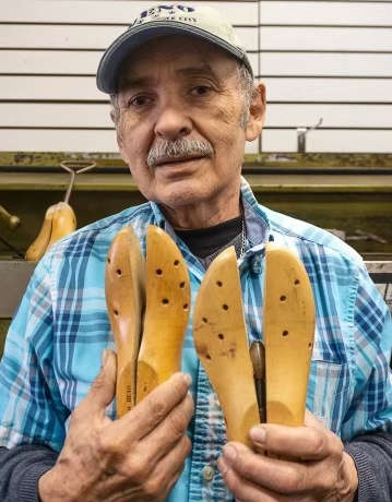 An older man in a plaid shirt and cap holds several wooden shoe stretchers in a workshop, with more tools and shoe stretchers hanging in the background.