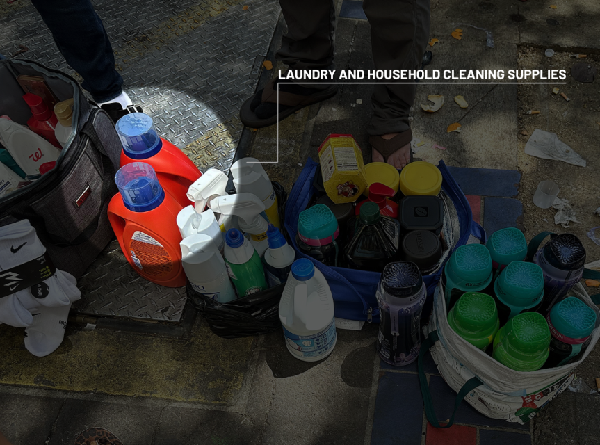 Bottles of laundry detergents and household cleaning products displayed on the ground next to people's feet outdoors.