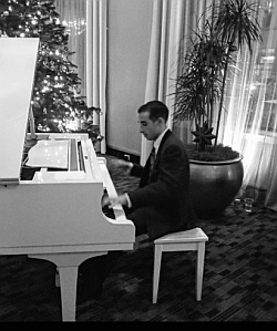 A man in a suit plays a white grand piano in a decorated room with a lit Christmas tree and potted plants.