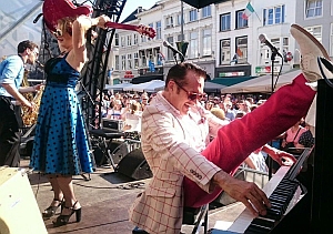 A man in a plaid jacket plays piano with one leg raised high, while a woman in a blue dress holds a guitar above her head on an outdoor stage before a crowd.
