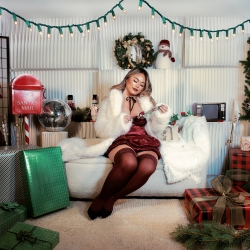 A woman in festive attire sits on a white couch surrounded by holiday decorations, wrapped gifts, and a mailbox, with string lights and wreaths in the background.