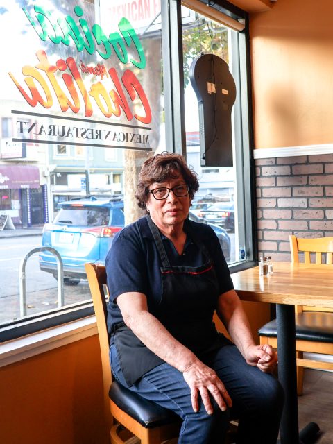 An older woman wearing glasses and an apron sits at a table inside a Mexican restaurant near a window with colorful signage.