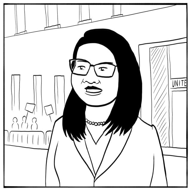 A woman with glasses stands outside a building, with people holding protest signs visible in the background.