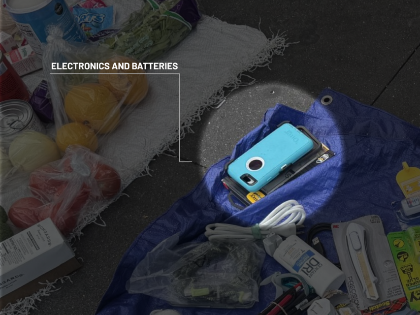 A blue smartphone with a case lies on a tarp among miscellaneous electronics, batteries, and groceries.