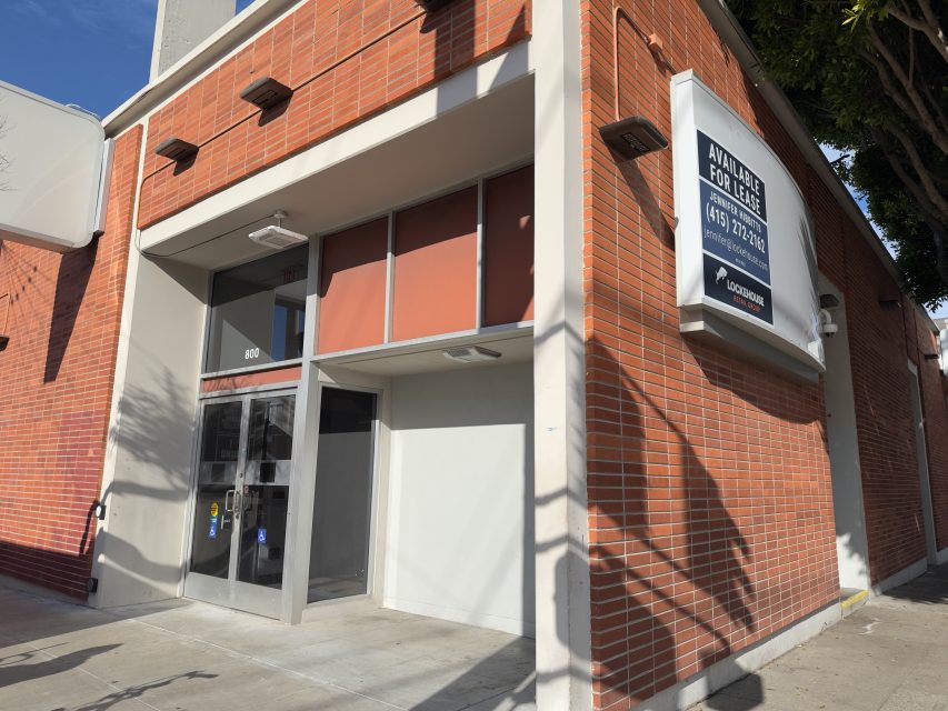A brick building with a glass door entrance and a large "Available for Lease" sign displaying a phone number on the exterior wall.