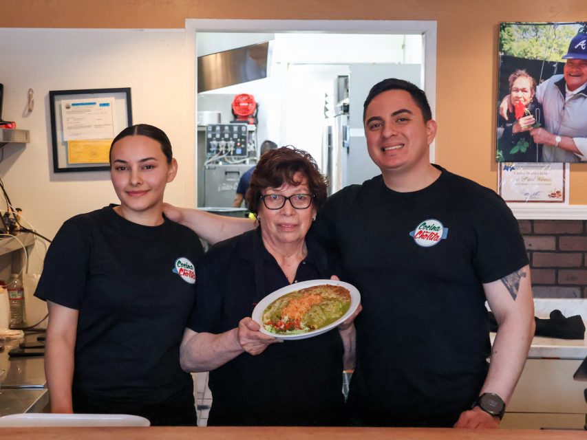Three people stand behind a restaurant counter; the woman in the center holds a plate of food while the others stand on either side, all wearing matching shirts.