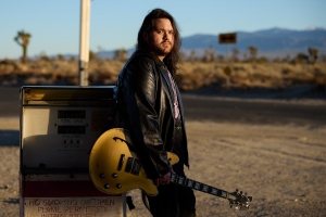 A man with long dark hair and a black leather jacket stands outdoors by an old gas pump, holding a yellow electric guitar. Desert landscape and mountains are visible in the background.