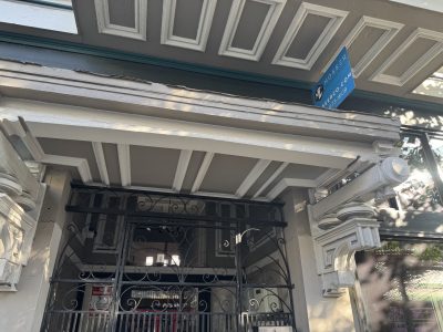 Ornate building entrance with decorative molding, black wrought iron gate, and a blue street sign displaying “Moss Pi” above the doorway.
