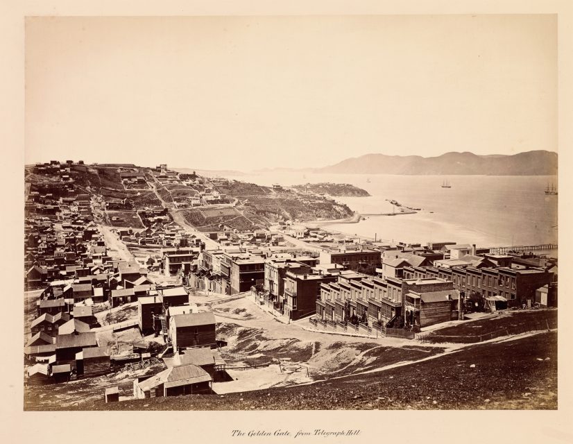 Black-and-white photograph of a coastal city with rows of wooden buildings, dirt roads, and hills, overlooking a bay with several ships in the water.