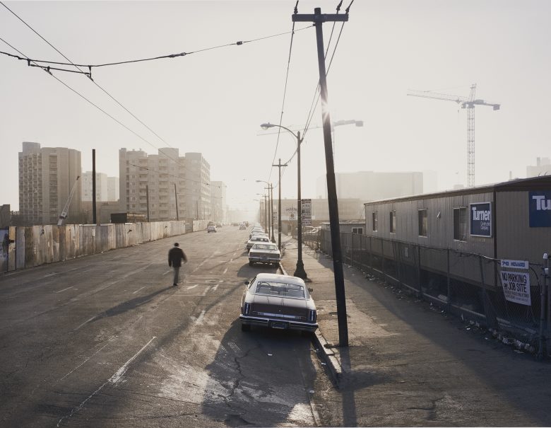A mostly empty urban street with parked cars, a lone person walking, construction fences, and cranes in the background under a hazy sky.