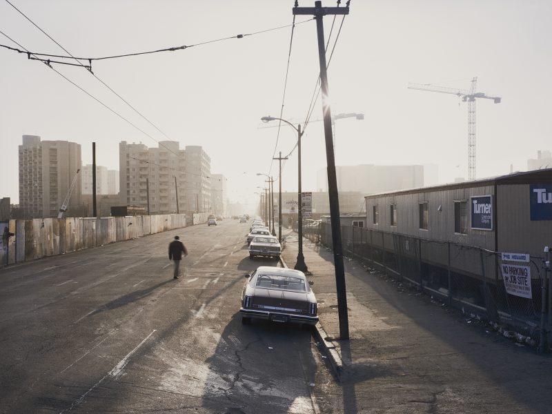 A mostly empty urban street with parked cars, a lone person walking, construction fences, and cranes in the background under a hazy sky.