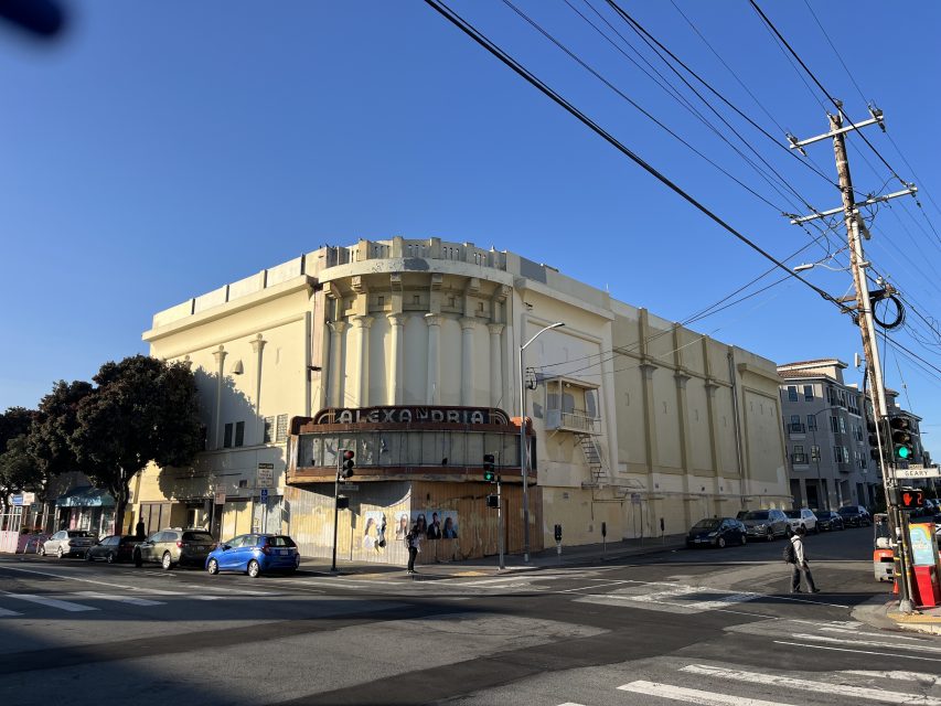 A large, faded yellow theater building with "Alexandria" on the marquee stands on a street corner, showing visible wear and graffiti. Cars and pedestrians are nearby under a clear blue sky.