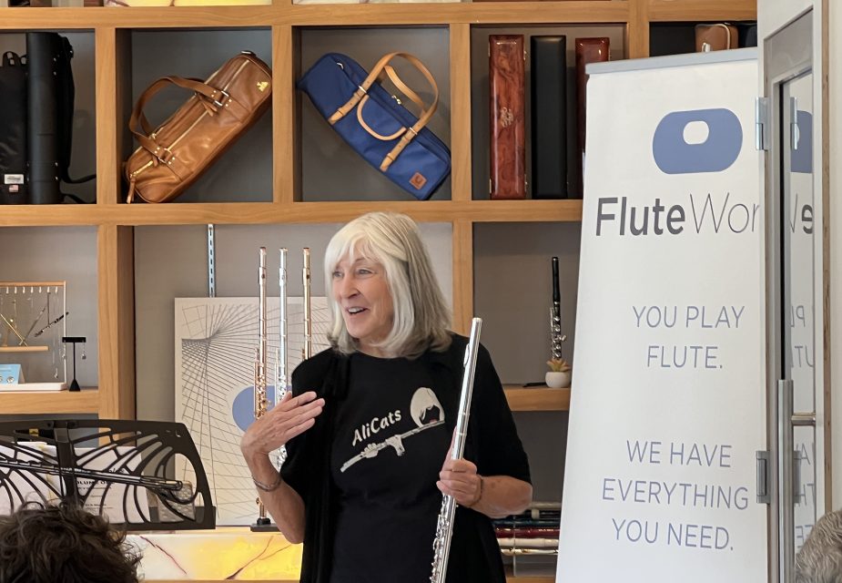 A woman holding a flute stands beside a "Flute World" sign, speaking to an audience in a room with shelves displaying flute cases and musical accessories.