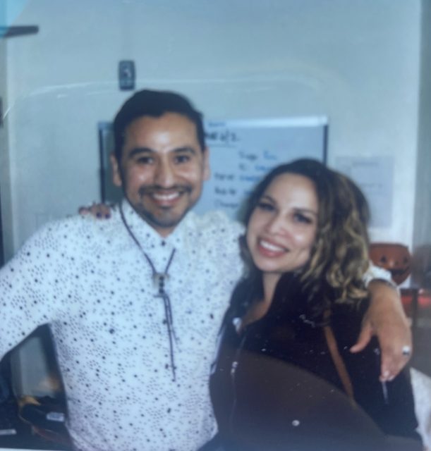 Two people are standing indoors, smiling at the camera. Alberto Rangel is wearing a white patterned shirt and the woman is in a dark outfit; a whiteboard is visible in the background.