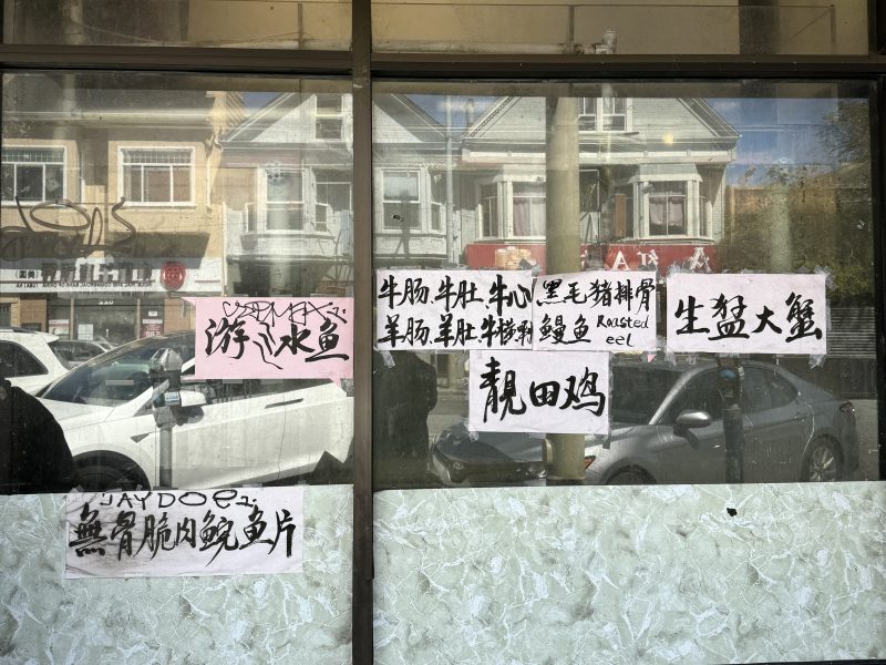 Chinese handwritten signs displayed on a storefront window, listing various food items; parked cars and residential buildings are reflected in the glass.