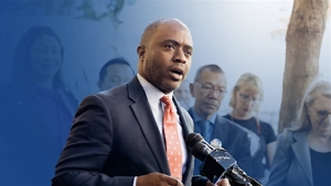 A man in a suit and tie speaks at a podium with a microphone, with several people standing in the background.