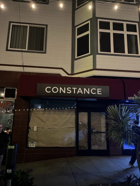 A dark street scene showing a storefront with a red awning and the word "CONSTANCE" in white letters; windows are covered with brown paper.