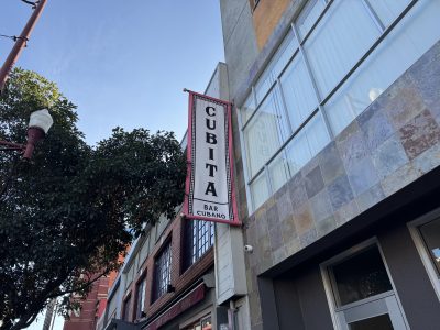 A vertical sign on a building facade reads "Cubita Bar Cubano" against a background of trees and a clear sky.