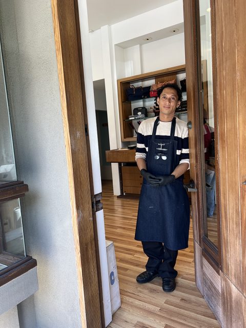 A person wearing a striped shirt, denim apron, and black gloves stands in the doorway of a shop with wooden floors and shelves in the background.