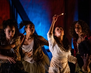 Four women in dramatic costumes stand in dim lighting, with one woman in white holding a lit match aloft, others looking on with intense expressions.
