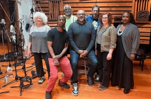Seven people pose together in a recording studio with microphones and soundproofing panels visible in the background.