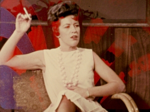Woman in a white sleeveless dress sits on a chair, holding a cigarette in her raised hand, with a wood-paneled background.