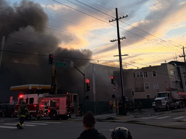 Firefighters respond to a building fire with heavy smoke billowing, fire truck ladders extended, and traffic lights visible at an urban intersection during sunset.
